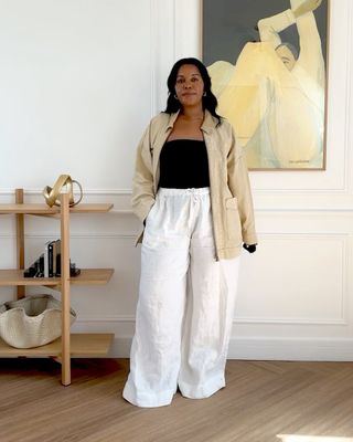 fashion influencer Style Idealist poses in a stylish room with wood shelves, modern decor, and an abstract painting, wearing a tan chore barn coat a black top, and white wide-leg linen pants