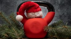 Red kettlebell with a red Santa hat resting on top of the bell, surrounded by pine branches