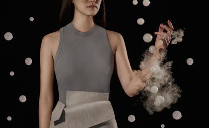 View of a woman in a grey sleeveless outfit surrounded by white bubbles and puffs of smoke in a space with a black background