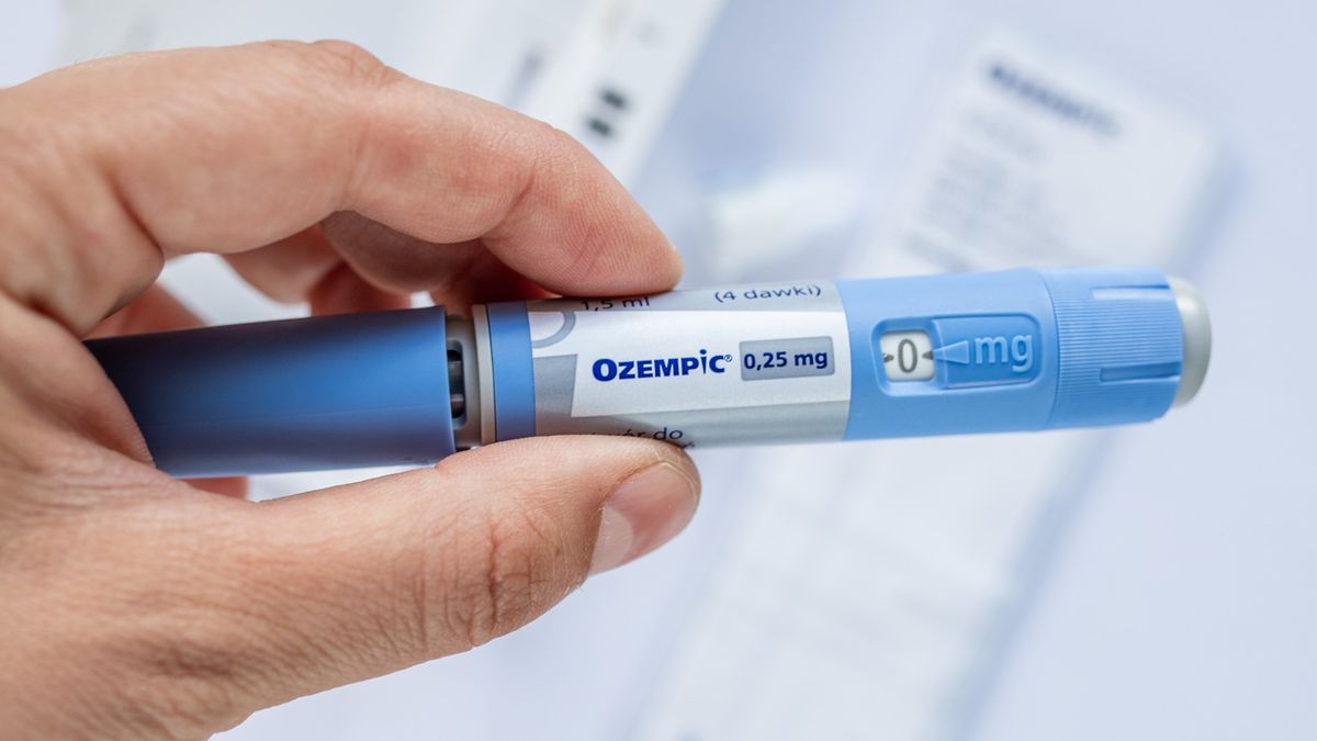 Close up of hand holding Ozempic injection. It looks like a thick, light blue, pen with &#039;Ozempic&#039; on the white label in dark blue writing. 