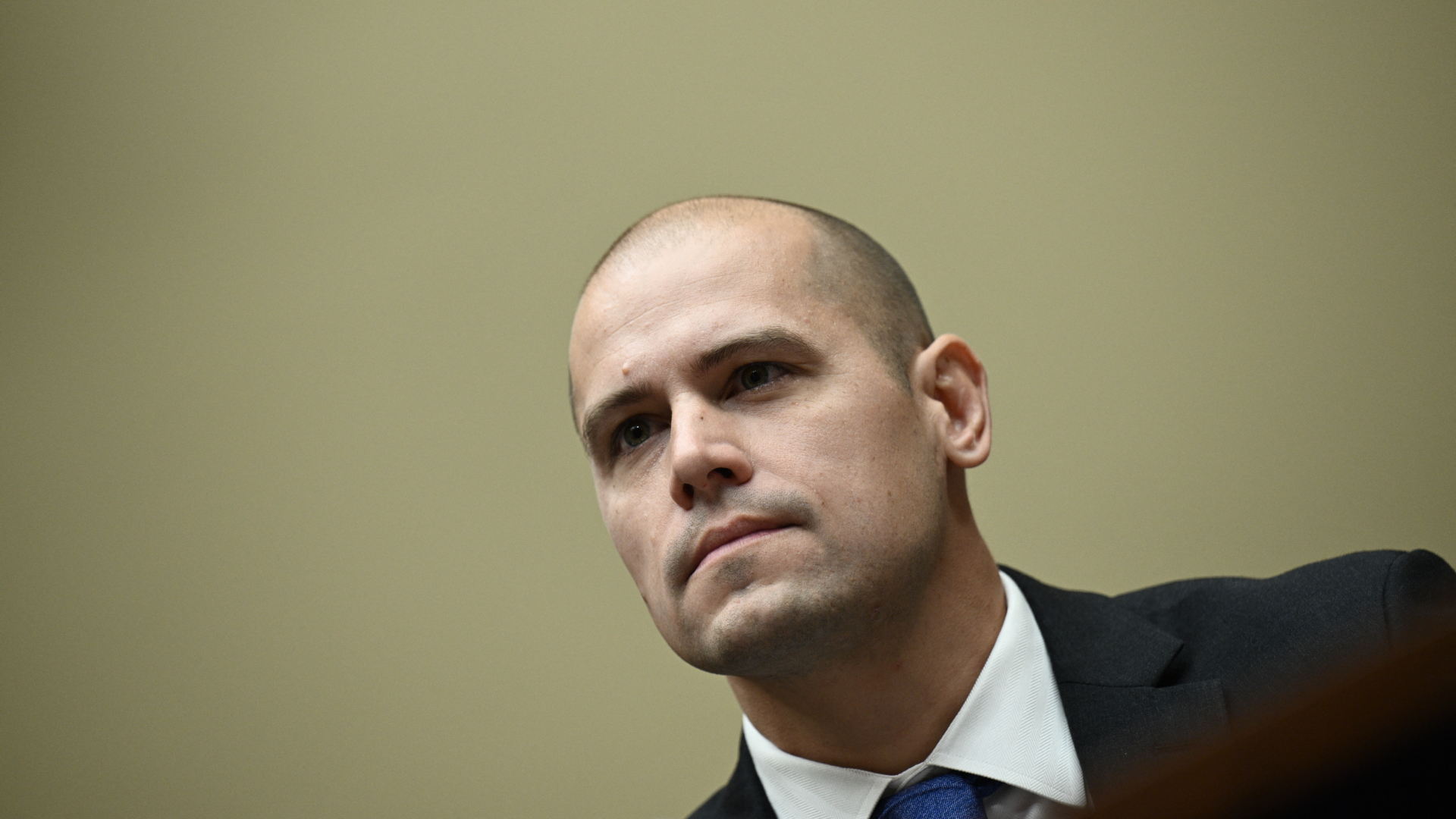 A bald man with a black suit and blue tie stands in front of a beige background and is seen in the bottom right of the image