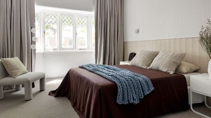 A bright bedroom with a boucle chair and cushion, a large bed with beige pillows and cushions, a plum duvet, a blue throw and a white side table