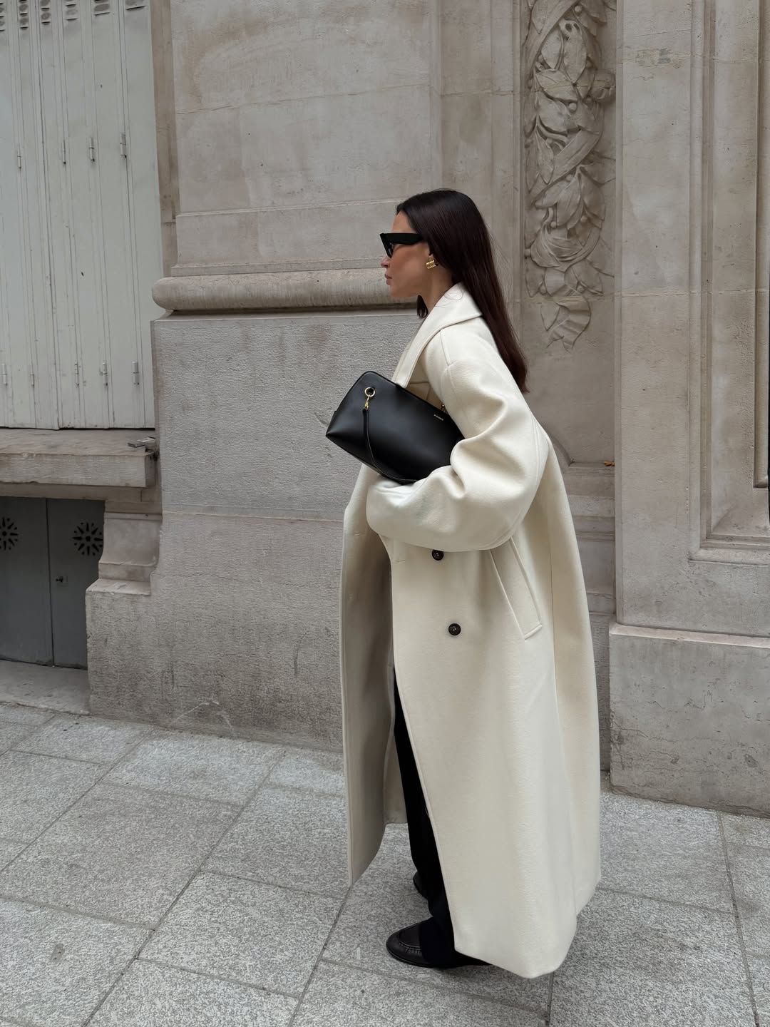 A woman wears a tan double-breasted coat with black pants and black leather boots.