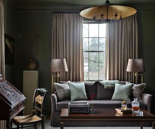 dark living room with dark green walls, gray sofa with cushions, dark wooden coffee table and writing desk with vintage chair