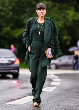 Katya Tolstova wears gold earrings, a god long chain pendant necklace, a khaki ribbed halter neck tank-top, a dark green long blazer jacket with embroidered feathers back details, matching dark green suit pants, gold shiny varnished leather platform heels shoes, a pink / purple / white embossed painted pattern clutch in shape of book, outside Saks Potts, during the Copenhagen Fashion Week Spring/Summer 2024 on August 07, 2023 in Copenhagen, Denmark.