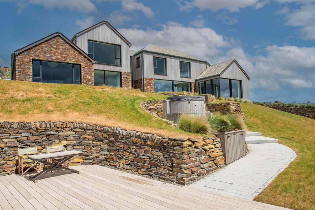 A walkway leading up to a brick house with large windows and hot tub and stone wall in front