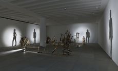 Wide view of the ’Nihilistic Optimistic’ exhibition. Shadow sculptures made out of junk, metal, and wood are placed throughout the area. Light shines on the sculptures and casts a shadow on a wall