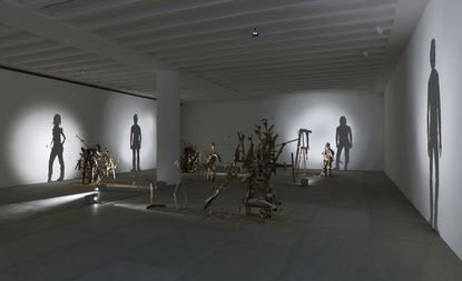 Wide view of the ’Nihilistic Optimistic’ exhibition. Shadow sculptures made out of junk, metal, and wood are placed throughout the area. Light shines on the sculptures and casts a shadow on a wall