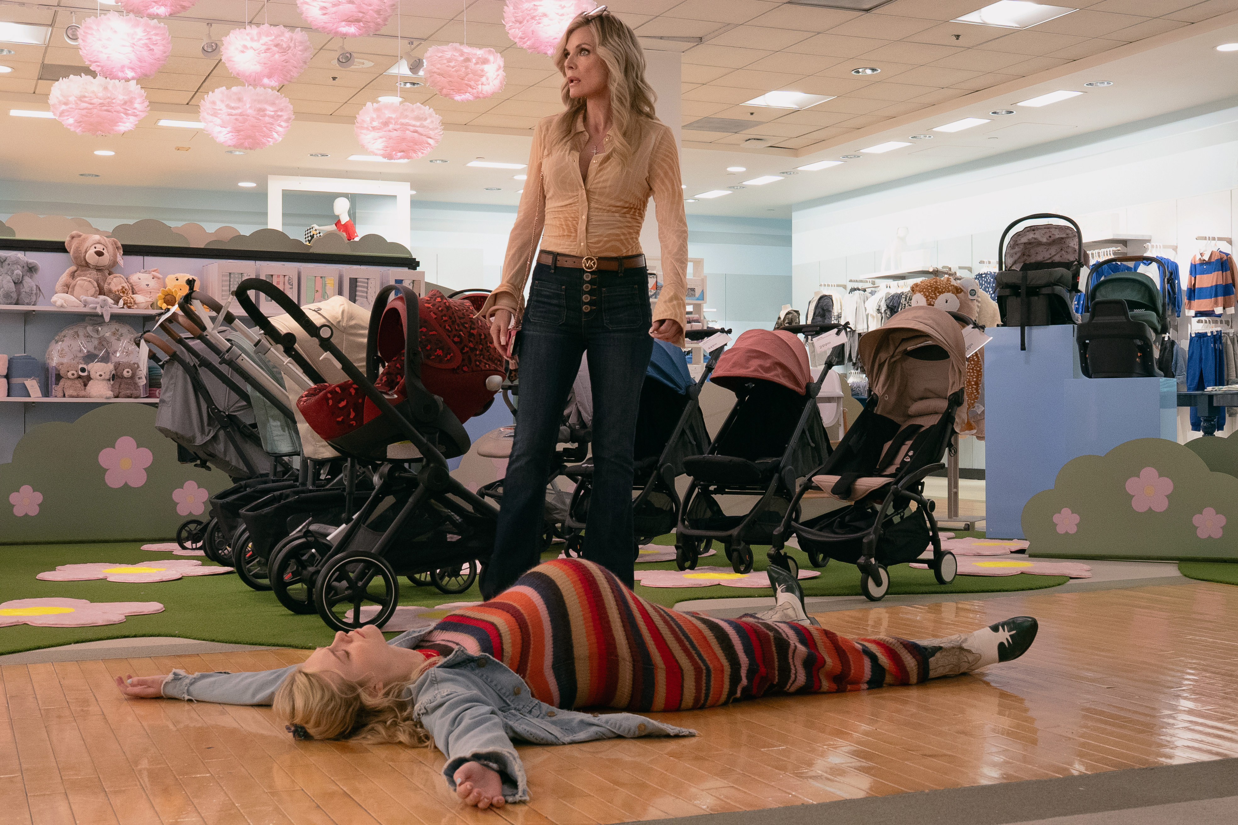 A blonde mother (Michelle Pfeiffer) stands over her pregnant daughter (Elle Fanning), who wears a striped sweater as she lies spread out on the wooden floor or a baby-clothing retailer, in Apple TV+'s 'Margo's Got Money Troubles.'