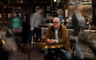 Paddy Kirk sits on a table with blurred people in the background in Emmerdale 