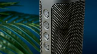 a close up picture of the three buttons of a black oblong-shaped bluetooth speaker with mesh covering and a carry strap photographed on a beige table in front of a navy wall