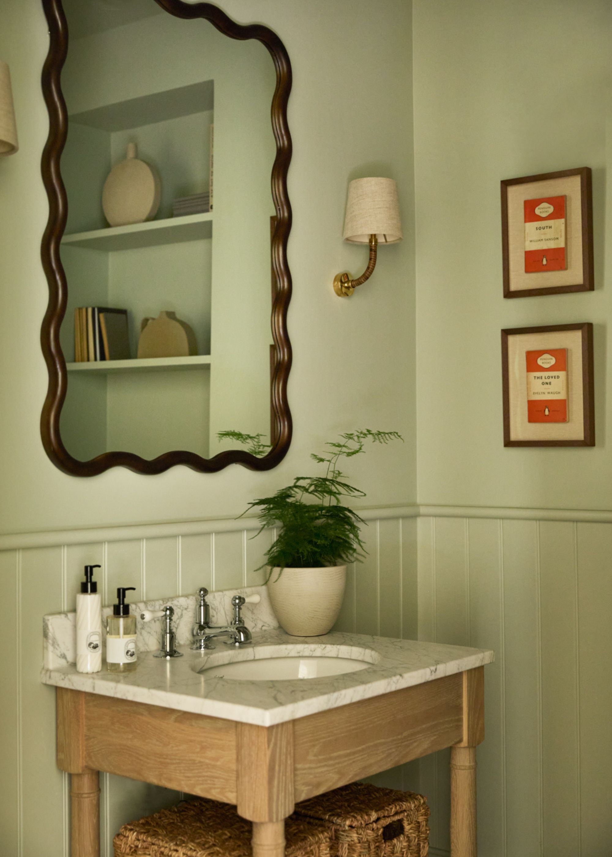 A small bathroom with soft green color-drenched walls with paneling on the lower half. A wooden sink vanity with a marble counter and a wavy mirror above it.
