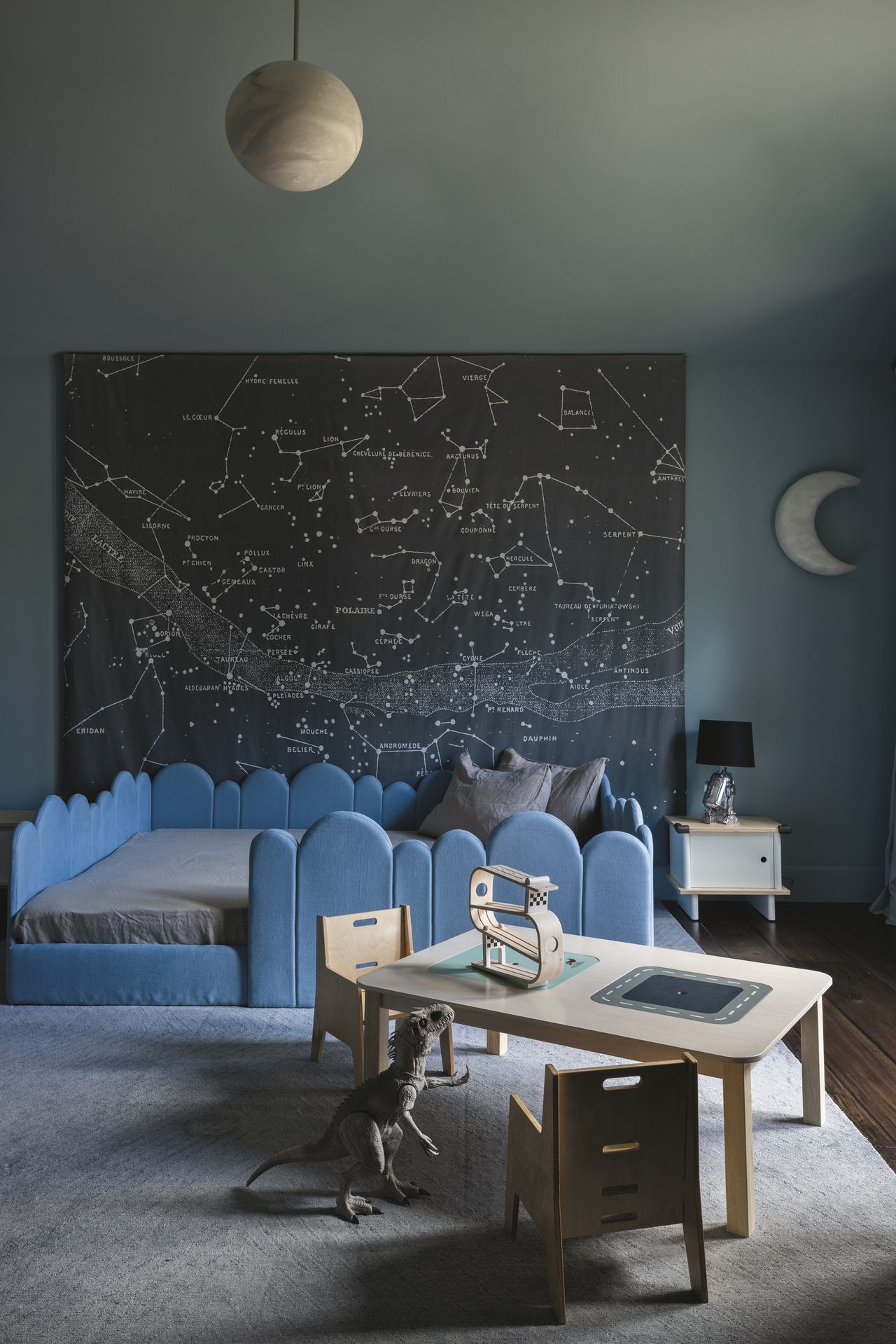 kids room with blue scalloped bedframe, blue walls and ceiling, moon-shaped pendant light, gray rug, dark timber floorboards, desk with two chairs, dinosaur toy, moon-shaped wall decal, and map of constellations hanging on the wall