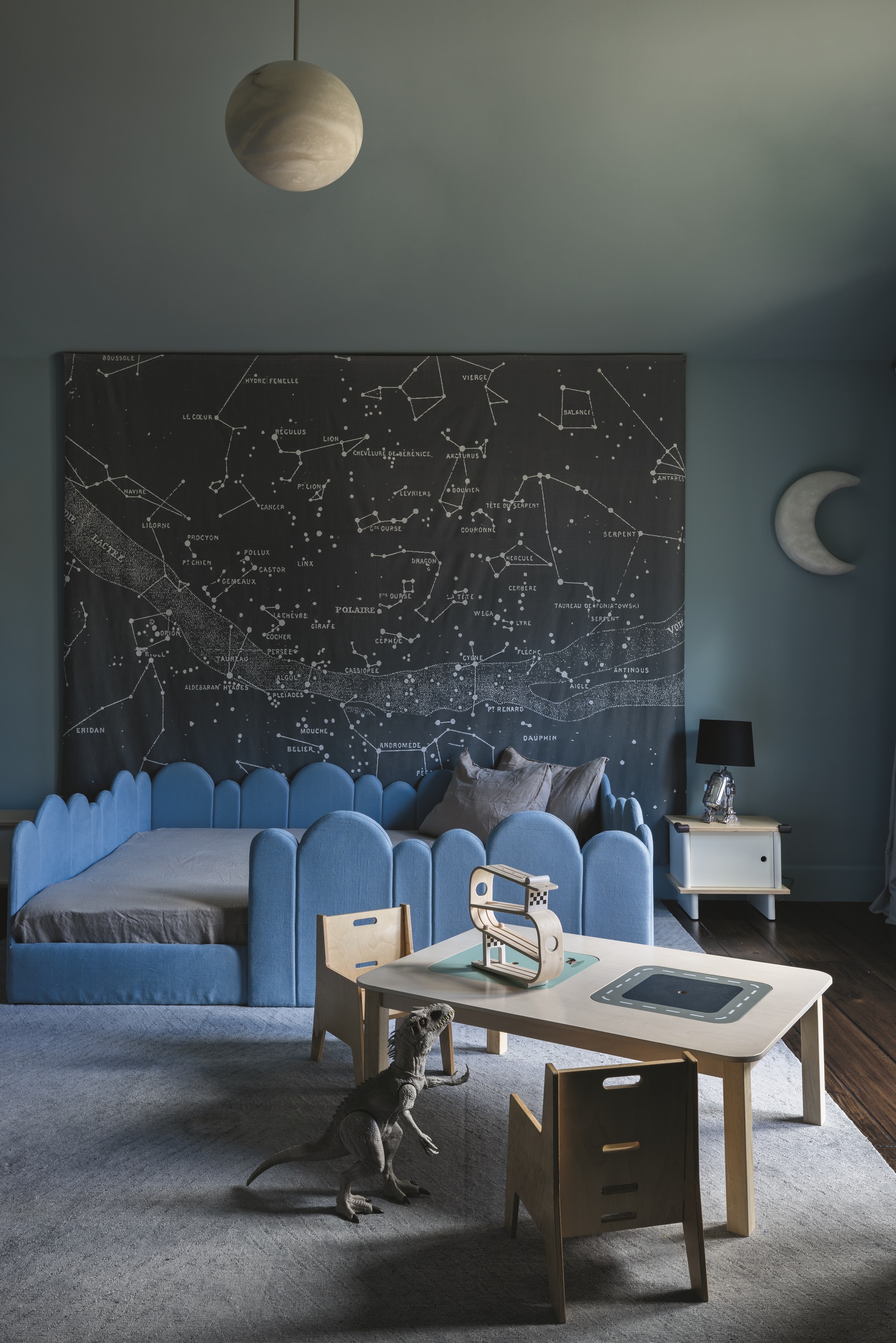 kids room with blue scalloped bedframe, blue walls and ceiling, moon-shaped pendant light, gray rug, dark timber floorboards, desk with two chairs, dinosaur toy, moon-shaped wall decal, and map of constellations hanging on the wall