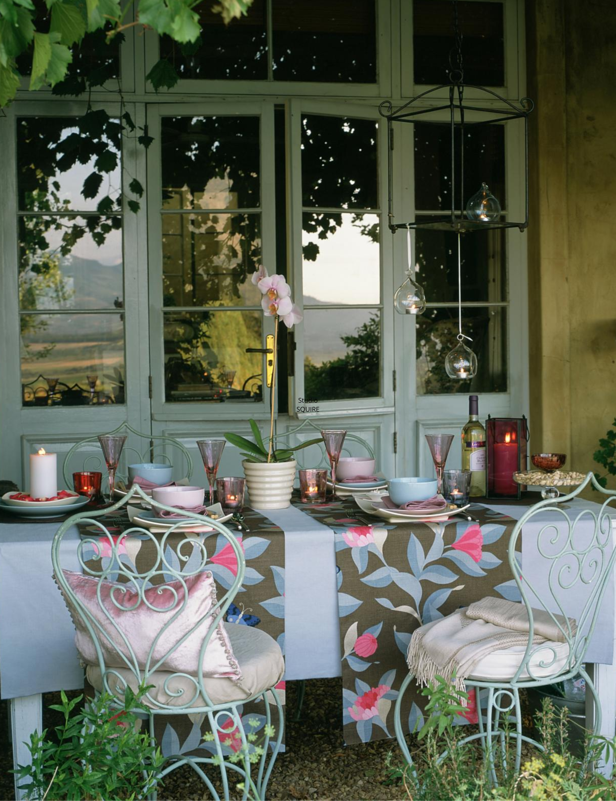 Whimsical outdoor tablescape featuring a sky-blue tablecloth layered with floral runner rose, rose-tinted wine glasses, and glowing candles.