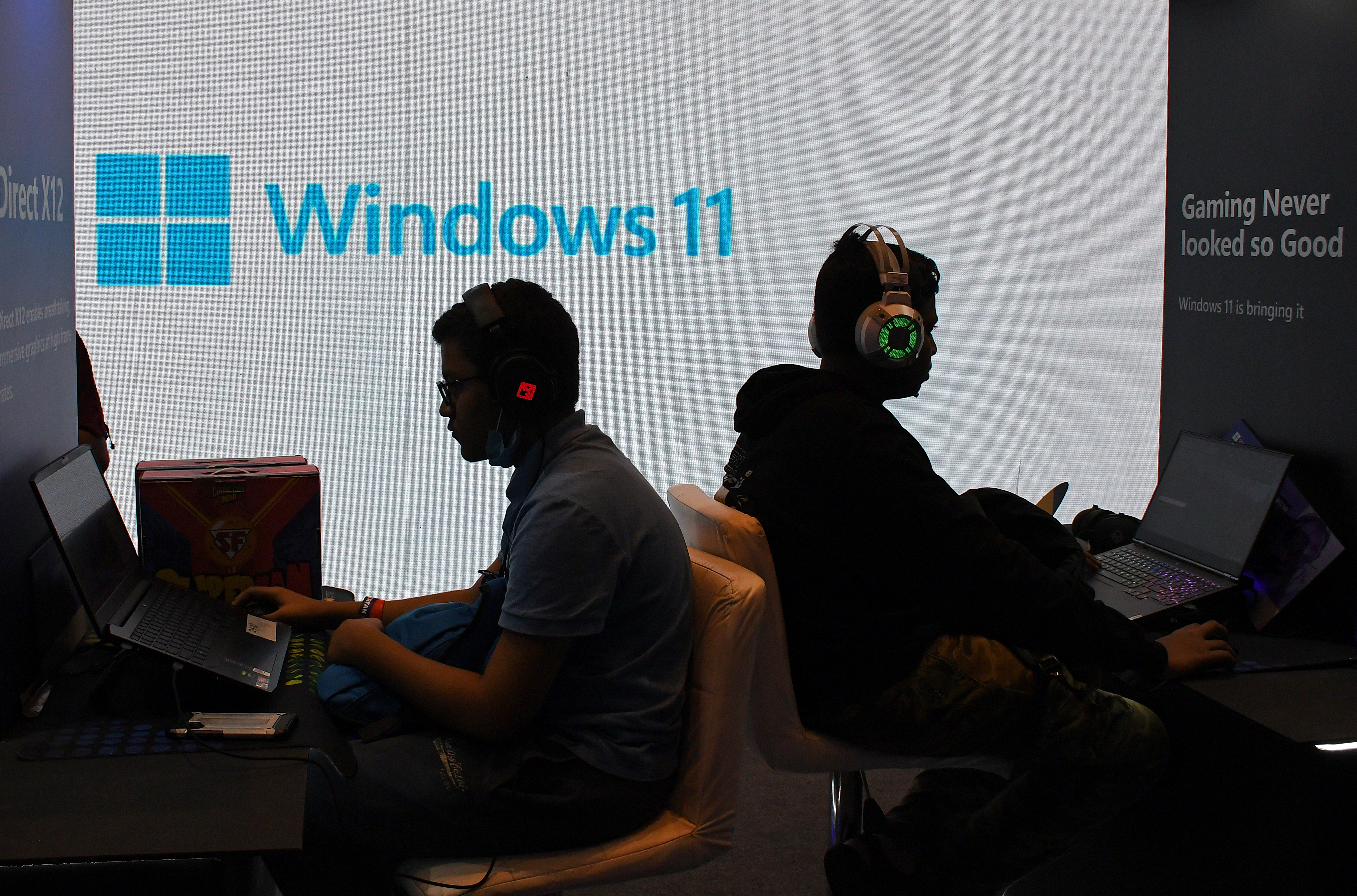 A Windows 11 logo looms in the background while two people wearing headsets with at computers with their backs to each other.
