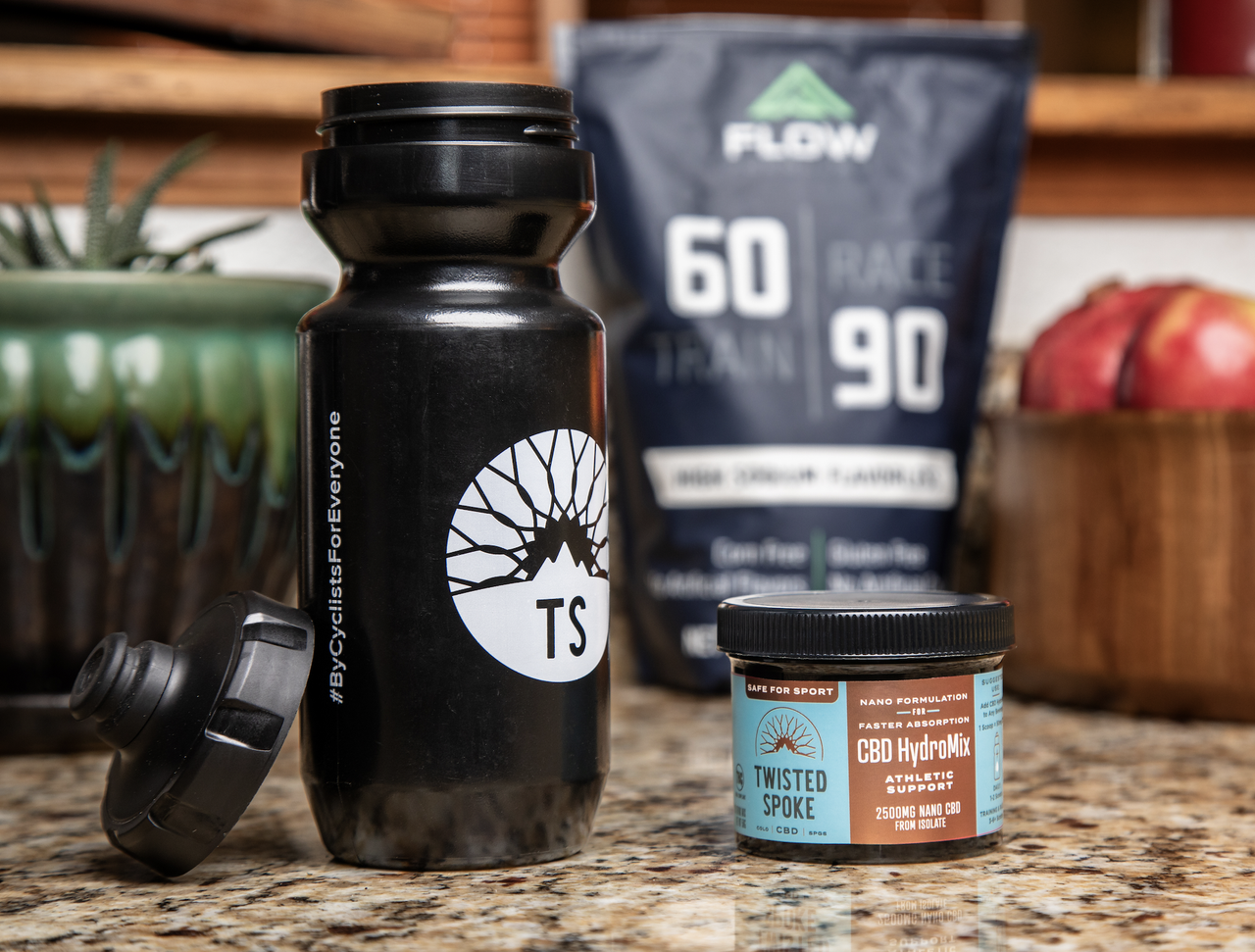 A black 20-ounce water bottle sits on the left and to its right is a 2500mg turquoise and bronze container of CBD powder. Both sit atop a mottled stone counter. In the background, there is a blurry bag of drink mix and a bowl of pomegranates. 