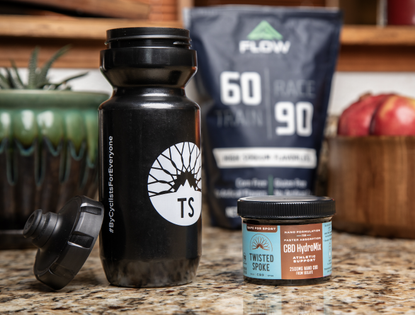A black 20-ounce water bottle sits on the left and to its right is a 2500mg turquoise and brown container of CBD powder. Both sit atop a mottled stone counter. In the background, there is a blurry bag of drink mix and a bowl of pomegranates. 