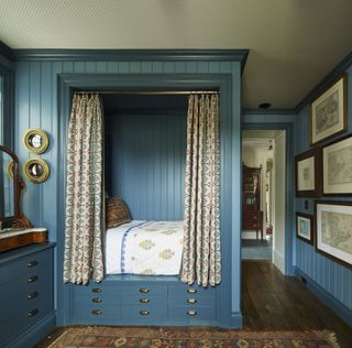 A blue sleeping berth surrounded up beaded board and behind a patterned curtain