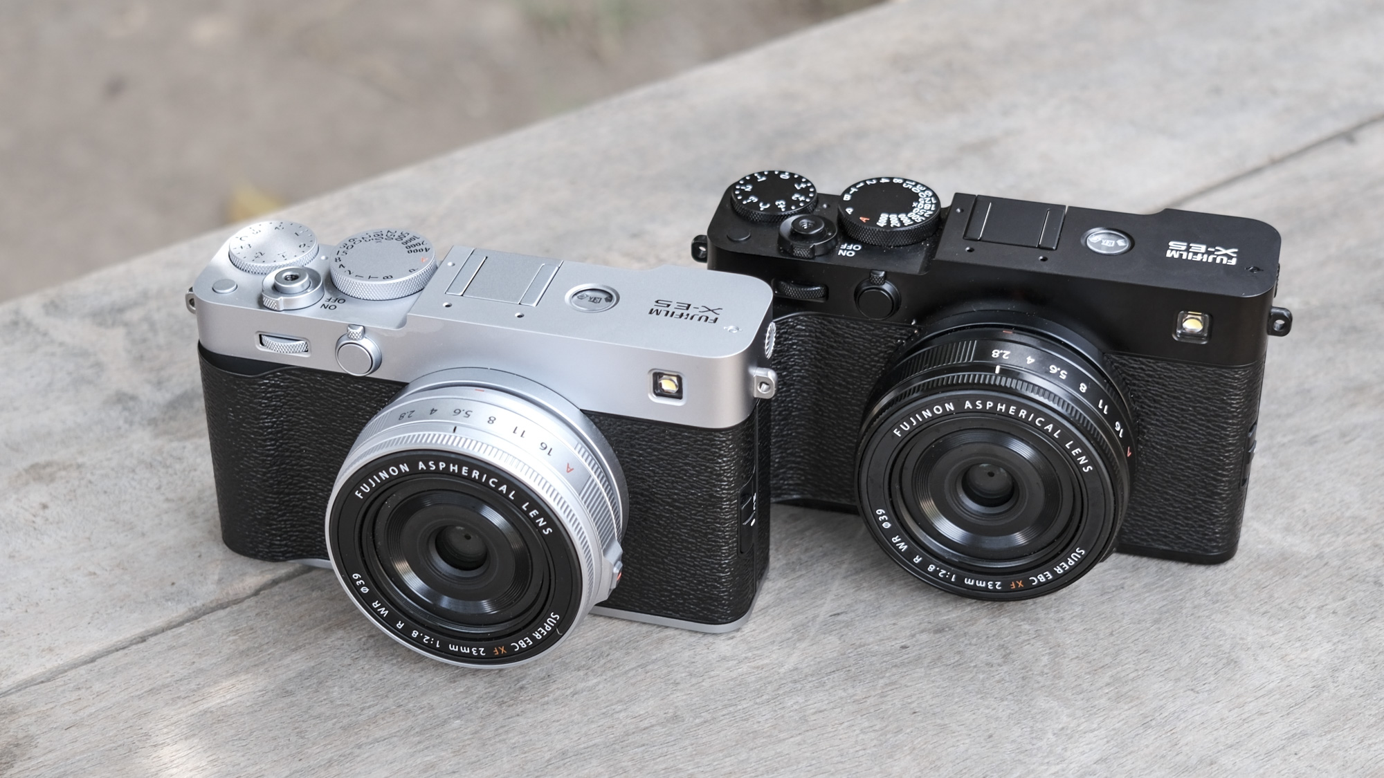 Black Fujifilm X-E5 alongside a silver Fujifilm X-E5, on a wooden bench