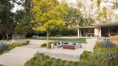 A large garden with a walkway and a mid point with built in seating and a fire pit in the middle. Beside the seating is a large bright green/yellow tree. There is also a view of the house