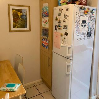 White fridge / freezer combination with various fridge magnets on it next to wooden cupboard on tiled floor