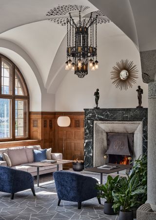 A sun-lit hotel lounge features mid-century velvet armchairs, sofas, and standing lights as well as plants within an Art Nouveau setting.