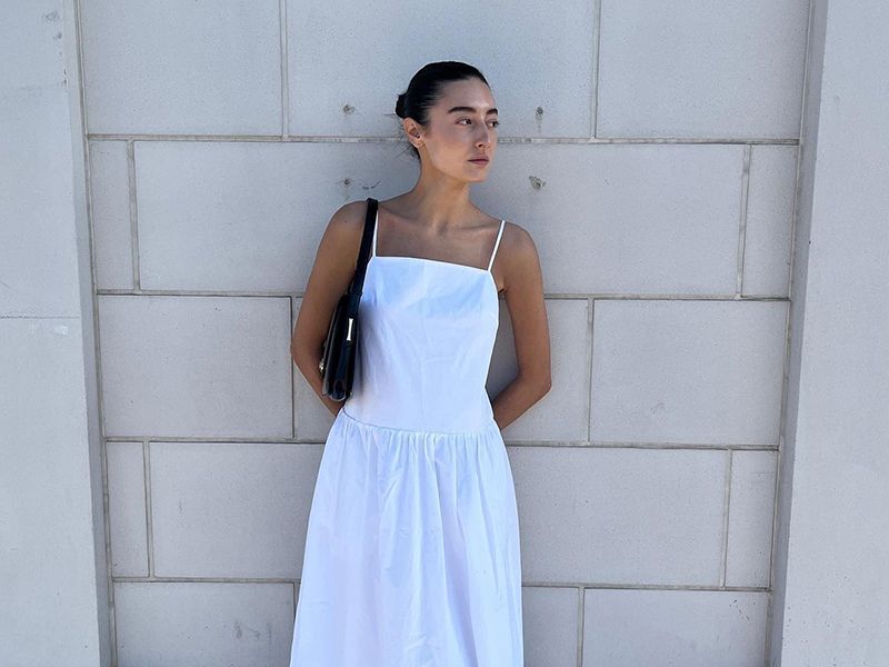 Dominique wears white linen dress with black shoulder bag and black mesh flats while leaning on a wall and posing.