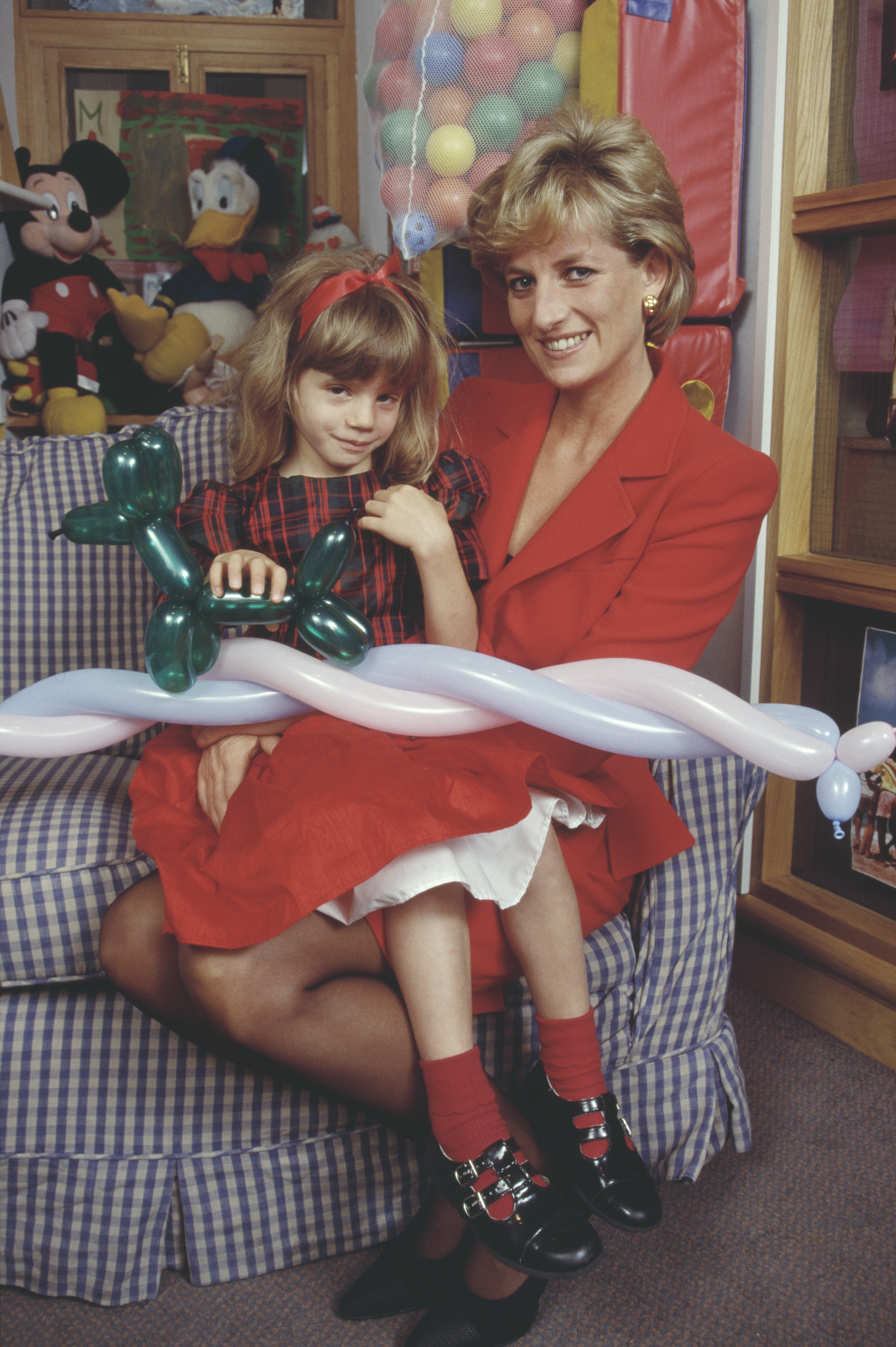 Princess Diana sitting on a gingham chair wearing a red skirt suit holding a little girl who is holding a balloon animal