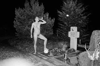 , Teller is stood in the nude near his father’s grave. He’s smoking, drinking, and balancing on a football.