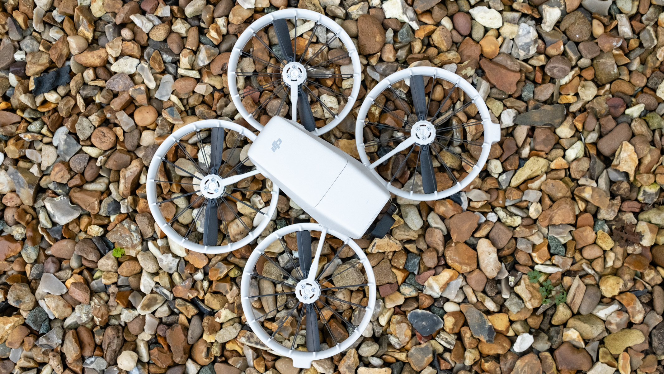 A white drone with four propellers sits on a stoney floor as we look down on it from a bird's eye view.