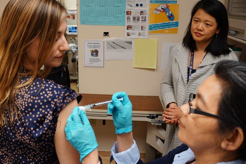 a photo of a doctor providing a vaccine to a young woman, a clinical trial participant, while a second woman looks on in the background