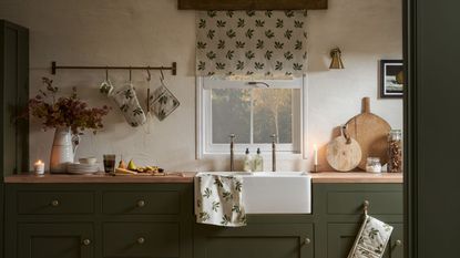 A kitchen with a large white farmhouse sink, dark green cabinets and light wood work surfaces. 