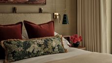 A bedroom with beige-gold texture wallpaper, a beige bed frame, and a bed styled with two red cushions, a floral bolster pillow, and a styled nightstand