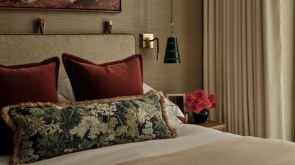 A bedroom with beige-gold texture wallpaper, a beige bed frame, and a bed styled with two red cushions, a floral bolster pillow, and a styled nightstand