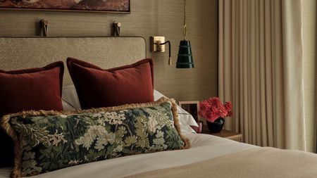 A bedroom with beige-gold texture wallpaper, a beige bed frame, and a bed styled with two red cushions, a floral bolster pillow, and a styled nightstand
