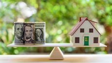 Rolls of cash sits on one end of a balance scale and a house on the other.