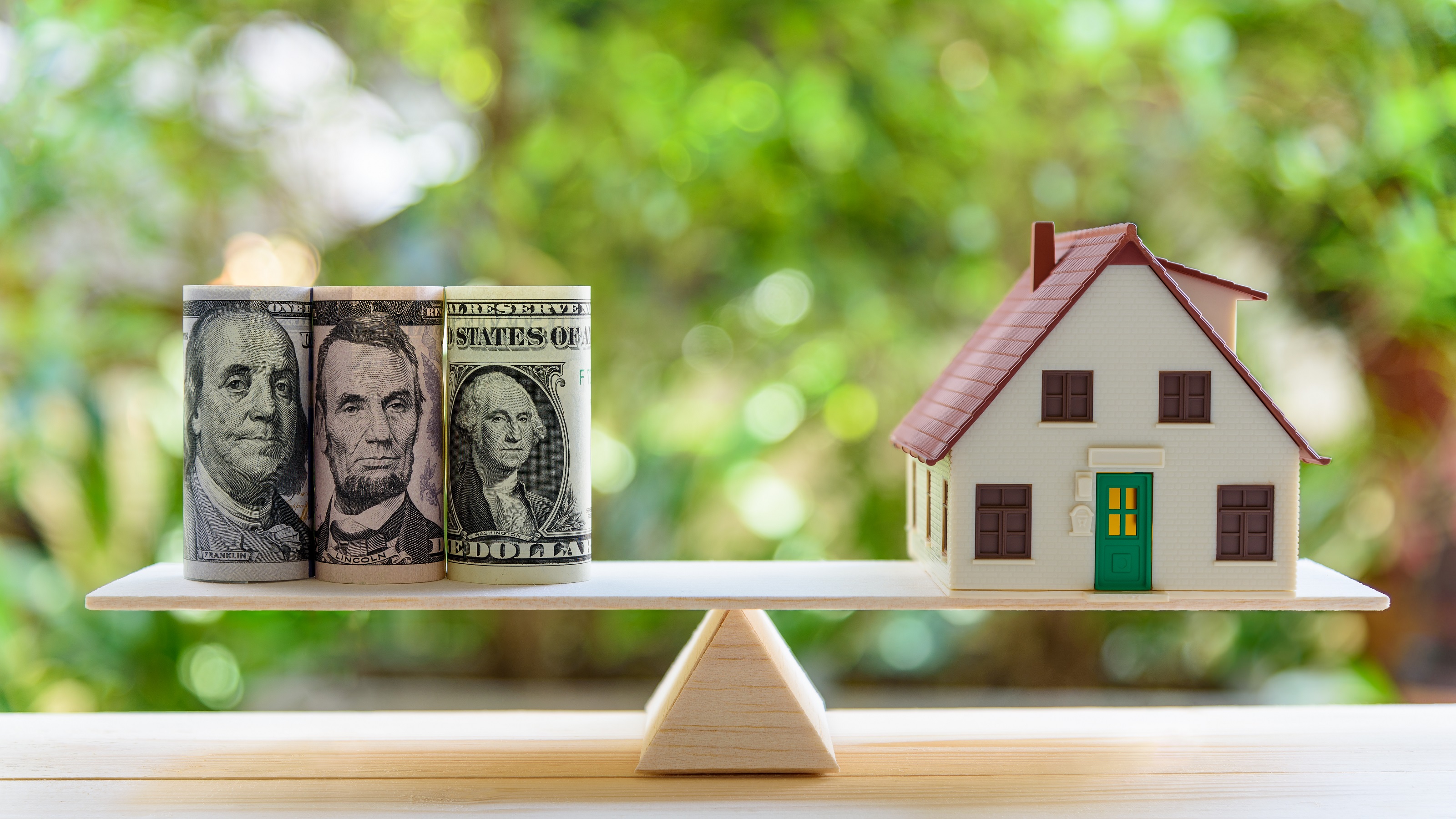 Rolls of cash sits on one end of a balance scale and a house on the other. 