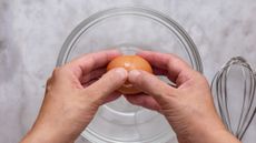 Two hands about to crack a brown egg into a glass bowl on a marble surface. A metal whisk sits to the right of the bowl.