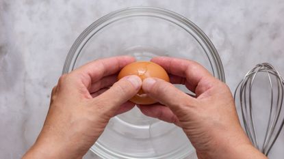 Two hands about to crack a brown egg into a glass bowl on a marble surface. A metal whisk sits to the right of the bowl.