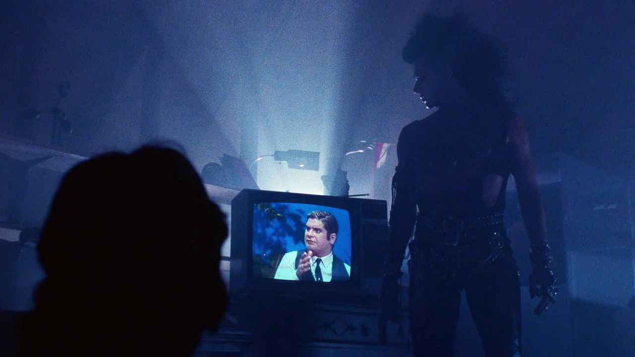 Tony Fields as Sammi Curr standing next to a TV with Ozzy Osbourne on it in Trick or Treat