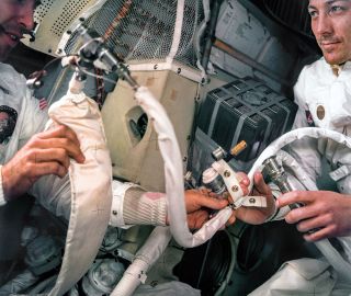 Apollo 13 astronauts Jim Lovell (at left) and Jack Swigert work with water hoses aboard the lunar module. A jerry-rigged carbon dioxide scrubber (or LiOH canister) is attached to the wall besides Swigert.