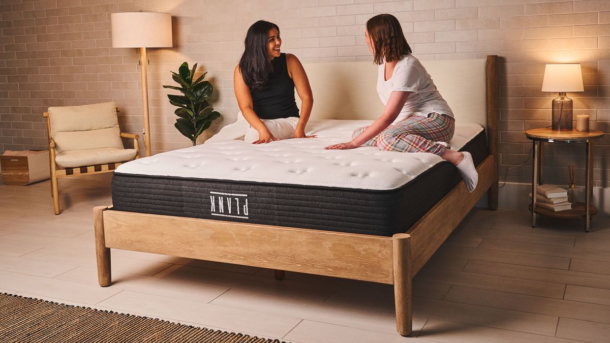Two women sit on the Plank Firm Mattress on a bed frame in a bedroom, discussing the feel and support