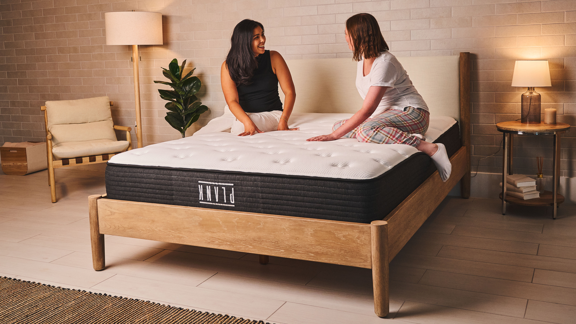 Two women sit on the Plank Firm Mattress on a bed frame in a bedroom, discussing the feel and support