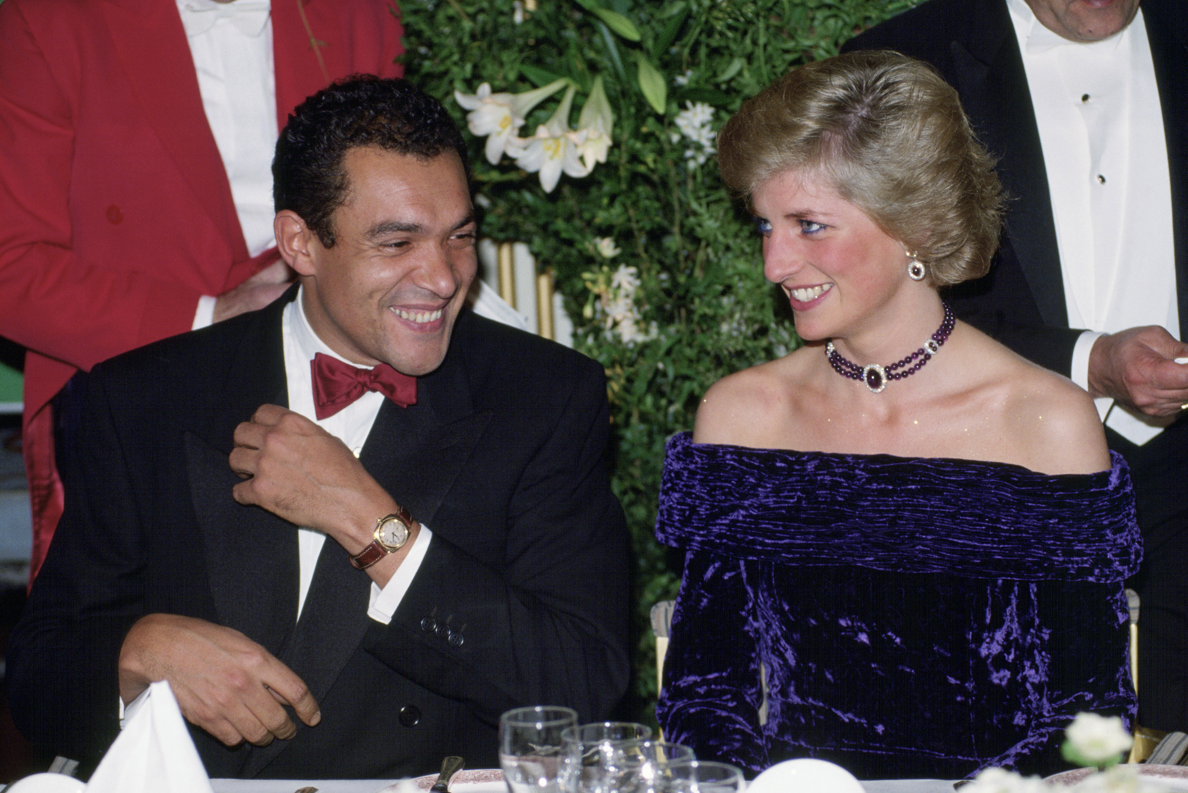 Princess Diana wearing an off the shoulder purple velvet dress sitting next to designer Bruce Oldfield