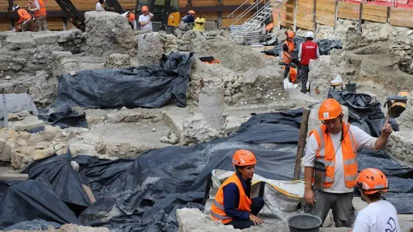about a dozen archaeologists in high-vis vests and hard hats work in an open excavation of low stone walls