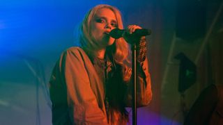 Sky Ferreira holding a microphone on stage with red light on her and blue lights on the background