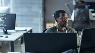 Male software developer using AI coding tools on a desktop computer while sitting in an open plan office space.