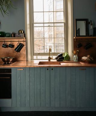 blue gray painted wooden kitchen with copper countertop and backsplash, shelf with rail underneath, cream painted window,