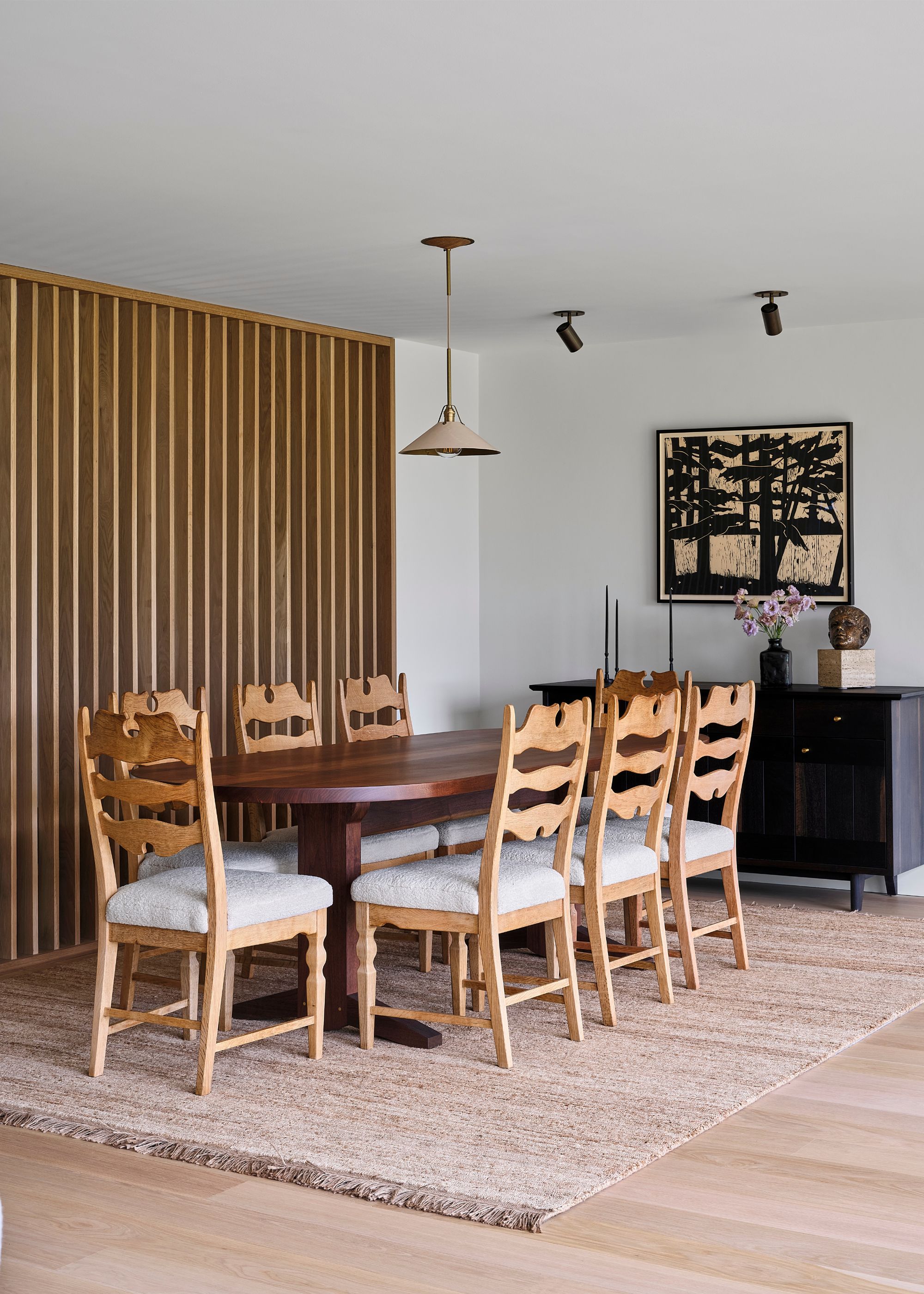 A modern dining room with white walls and a slatted wood divider wall. A beige textured rug and a large wooden dining table, and chairs.
